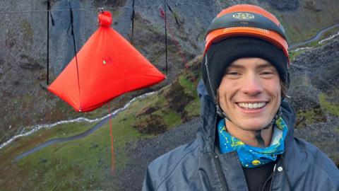 Joel smiles in front of a tent that is suspended in the air above a cliff face.