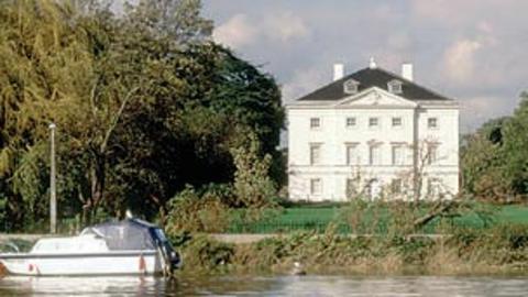 A large white villa set on the side of a large lake with two boats in the foreground.