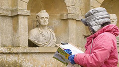 A young girl writing on a clipboard while looking at ornamental statue heads.