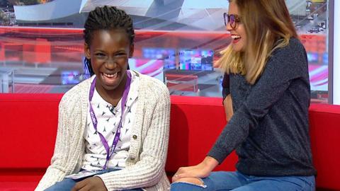 A girl looks excited and surprised, sat on a red BBC News sofa with a woman.