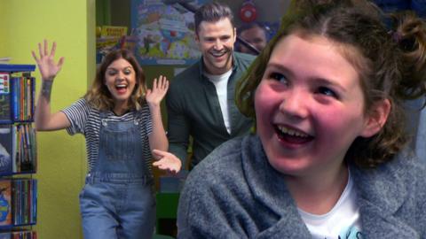 A girl looks excitedly on as two presenters jump out to surprise her in a library!