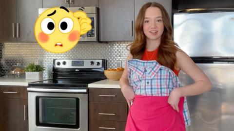 A young girl is in her kitchen looking directly into camera, she is holding a shirt and some fabric on her torso to form a dress. Beside her is a confused emoji scratching its head