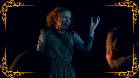 A Victorian girl holds up an item and smiles. From CBBC series Hetty Feather.