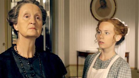 A girl in a victorian servant uniform looking up at an elderly lady who looks cross.
