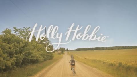 Holly Hobbie rides a bike down a country road, the series logo is overhead.