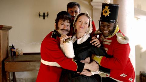 Florence Nightingale smiles while holding her lamp, Crimean soldiers laugh and smile with her hugging her.