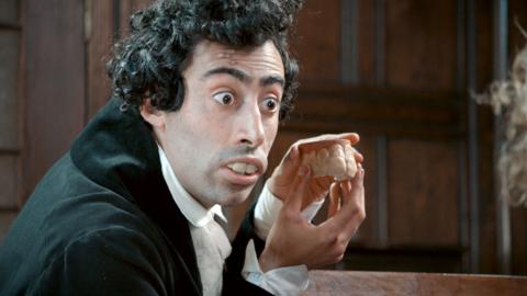 A man with very large teeth holds a set of dentures and looks wide-eyed at an unseen couple.