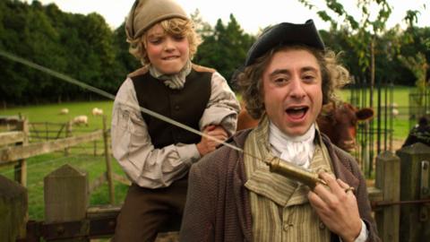 Horrible Histories cast member dressed up as Edward Jenner holding a large syringe as a boy dressed as James Phipps looks on nervously.