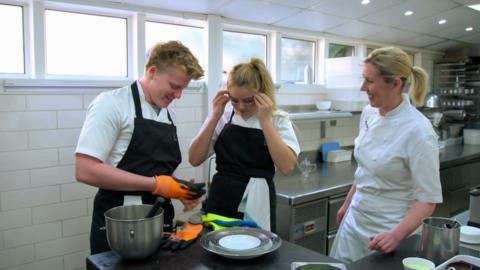 A boy and a girl put on safety goggles in a kitchen as a supervising chef smiles.