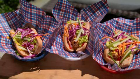 Three hot dogs covered with salad in American style fast food baskets sit on a table.