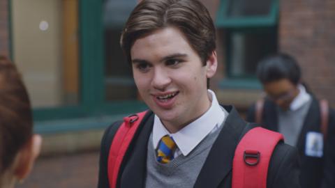 A teen boy wearing a school uniform and a red backpack smiles at a girl in the school playground. (Craig).
