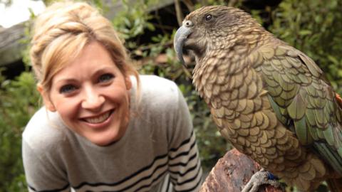 A woman leans next to a large green bird called a Kea on a branch. (Naomi).