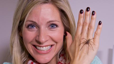 A woman's face smiling while holding her hand next to her face with a house spider sat across it (Naomi).