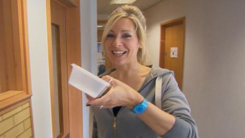 A woman with blonde hair stands in a school hallway looking towards the camera holding tupperware containing headlice (Naomi).