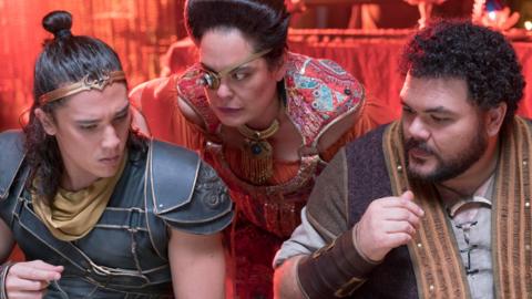 Three people in bright colours are sat at a table looking confused.