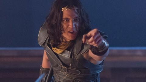 A man in armour with long hair and a golden crown, points angrily at someone.