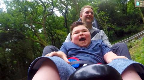 A man and a young boy on a rollercoaster in the woods.