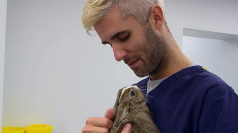 A man wearing blue scrubs holds a small rabbit against his chest.