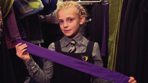 A girl wearing a school uniform holding a purple sash in front of her surrounded by costumes, Sybil from the Worst Witch.