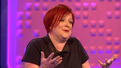 A woman with bright red hair shrugs her shoulders and smiles against a bright pink background.