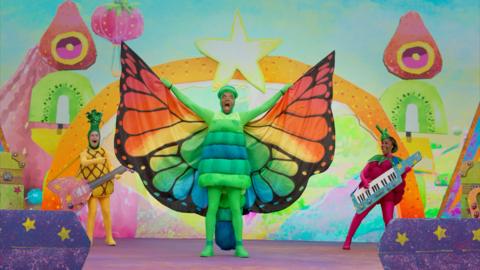 A butterfly, CBeebies presenter Rhys in a green work costume, with bright colourful wings.