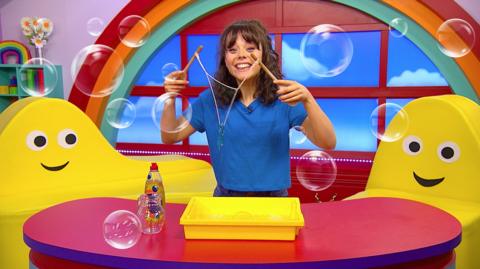 Evie is holding up her string bubble wand in CBeebies House, surrounded by bubbles.