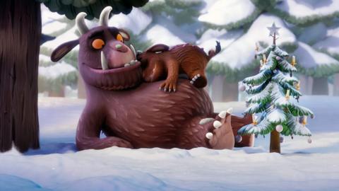 The gruffalo holding his child in the snow, next to a decorated Christmas tree.
