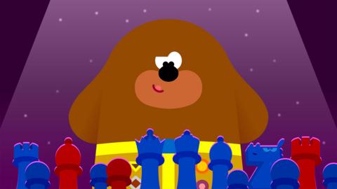 Duggee, a large brown cartoon dog, peers over a chess board, looking confident.