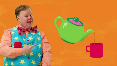 Mr Tumble is wearing a blue waistcoat with yellow stars. He is pointing at a tea pot pouring a drinking into a cup.