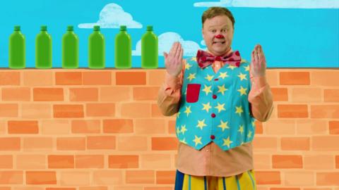 Mr Tumble is holding his hands up, there is a brick wall behind him with six green bottles on.