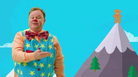 Mr Tumble is pointing towards an illustration of a bear standing at the top of a mountain.