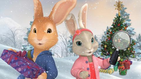 Peter Rabbit is holding a present and smiling to camera as Lily Bobtail holds a magnifying glass in front of a Christmas tree surrounded by presents. Behind them is a snowy background.