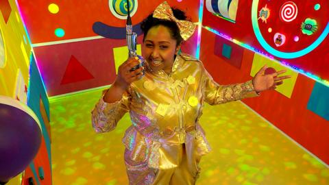 Bella stands in a gold jumpsuit speaking into a microphine in Playtime Towers lift.