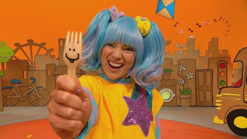 Sketch, a blue-haired woman, grins and holds up a wooden fork with a funny face on it.