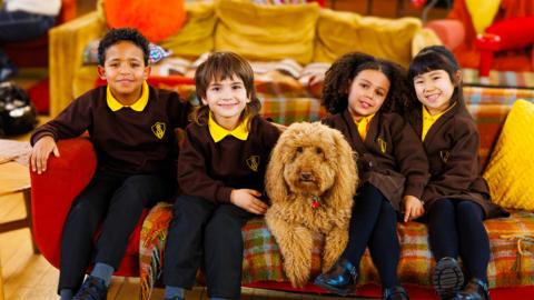 Four children in school uniform and a dog sit on a sofa, smiling.