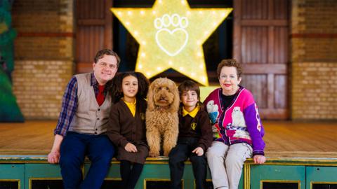A teacher in a brown vest, an older woman in a colourful cardigan, two children and a dog sit on a stage.