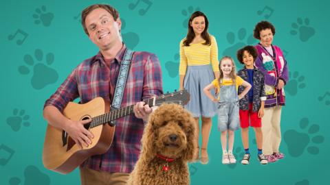 Simon is playing on his guitar with Waffle next to him, the rest of the family are stood behind smiling.