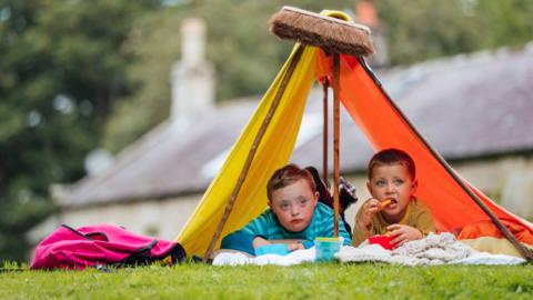 Two boys sit inside a makeshift tent built with blankets and a broom, most likely in their garden, watching the rain and eating cheese puffs.