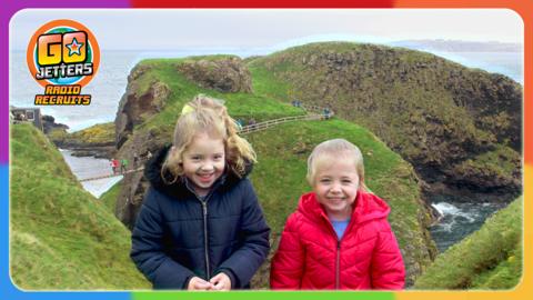 Go Jetters Radio Recruits Maggie and Iona are standing in front of a rope bridge with people crossing to Carrick-a-Rede.