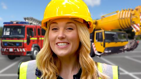 Maddie wearing a yellow safety helmet and high-vis jacket. Stood in front of a parked fire engine and crane.