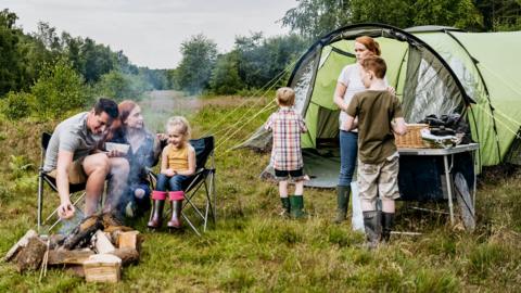 Family on a campsite, children gather around the tent and campfire with parents toasting marshmallows.