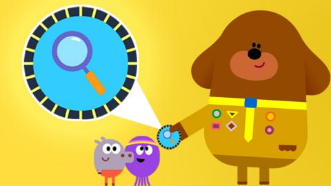 Hey duggee is handing the squirrels a badge. The badge is blue with a magnifying glass on it.
