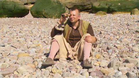 A pirate sat on a white stony beach, looking through a telescope