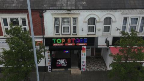 An aerial view of Top Store on Nantwich Road in Crewe. The shop is below a row of residential flats, with trees on the left and right of the shot. There is a multi-coloured sign advertising the shop with Tobacco Vape and Drinks beneath it.