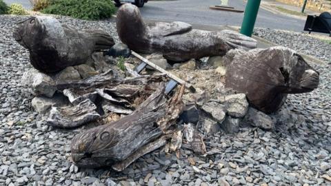 Three weathered brown seal sculptures surround the fragmented rotten remains of the unluckiest seal which has collapsed on itself. They are surrounded by slate chippings on a roundabout.