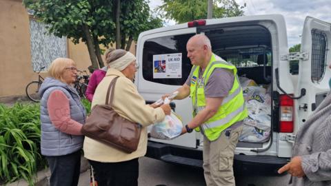 Gary Fear, a man wearing a high visibility jacket, is handing a white plastic shopping bag to a woman. There are two other women stood nearby. Behind them there is a mini van with one door open that contains more shopping bags.
