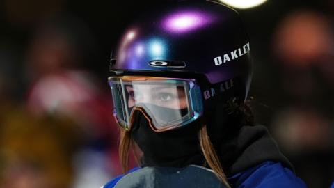 A woman wearing a helmet, goggles and a black face covering rests her chin on the edge of her snowboard.