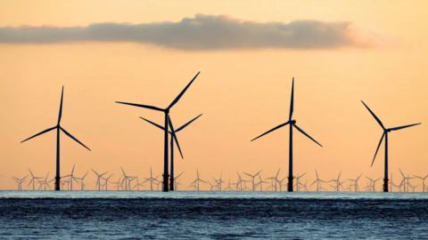 A large offshore wind farm with a number of turbines quite close, with tens of them in the distance, against an orange sky.