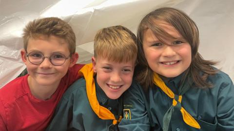 Three smiling boys aged around 11 side-by-side. Two are wearing scout uniforms with green and yellow neckerchiefs.