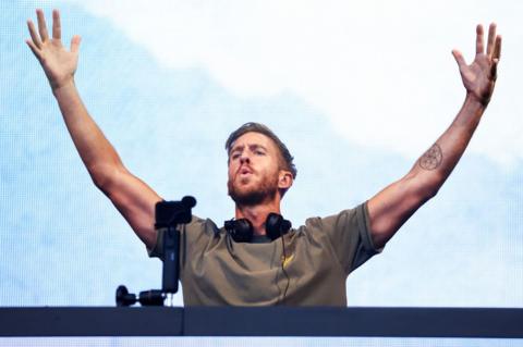 Calvin Harris with his arms in the air, behind DJ decks